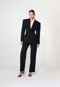 Femeie îmbrăcată într-un costum negru perfect croit, cu umeri puternici, bluză crem și sandale negre cu toc înalt, stând pe un fundal alb.