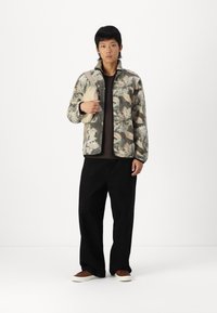 Fleece jacket with a camouflage pattern in shades of green and beige. Paired with a black shirt and wide-leg black pants, holding a beige bag.