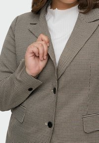 Žena upravující klopu hnědo-černého houndstooth blejzru, který má na sobě přes bílý žebrovaný top, zblízka na horní části těla a ruku.