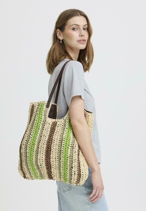 Giovane donna con maglietta grigia e jeans che porta una grande borsa a tracolla tessuta con strisce verticali verdi e marroni.