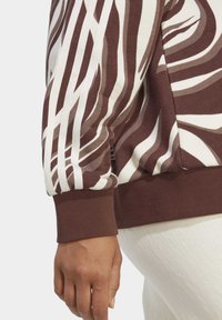 Sweatshirt listrado em castanho e branco com punhos canelados, apresentando um corte descontraído e tecido macio, mostrado de lado com uma mão pousada no quadril.