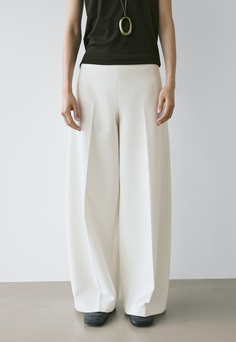 Femme portant un pantalon blanc à jambes larges, un haut noir sans manches, des chaussures noires à bouts carrés, et un long collier pendentif, debout devant un fond uni.