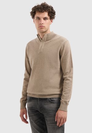 Mann mit lockigem Haar, der einen beigen Reißverschluss-Pullover und ausgewaschene schwarze Jeans trägt, steht vor einem einfarbigen, hellen Hintergrund.