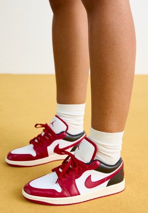 Nogi w białych skarpetkach i czerwono-biało-czarnych butach Nike Air Jordan stojące na żółtej podłodze.