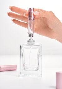 Przezroczysta szklana butelka perfum o prostokątnym kształcie, z srebrnymi elementami i różowym rozpylaczem, trzymana częściowo przez rękę.