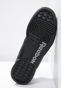 Černá gumová podrážka tenisky Reebok, se strukturou grip vzory a výrazným logem v bílé. Čistý, minimalistický design.