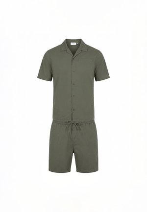 Mand stående i olivengrøn kortærmet skjorte med knapper og matchende shorts med sorte slip-on sko på hvid baggrund.
