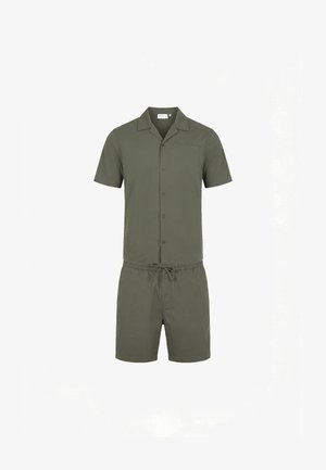 Mand stående i olivengrøn kortærmet skjorte med knapper og matchende shorts med sorte slip-on sko på hvid baggrund.