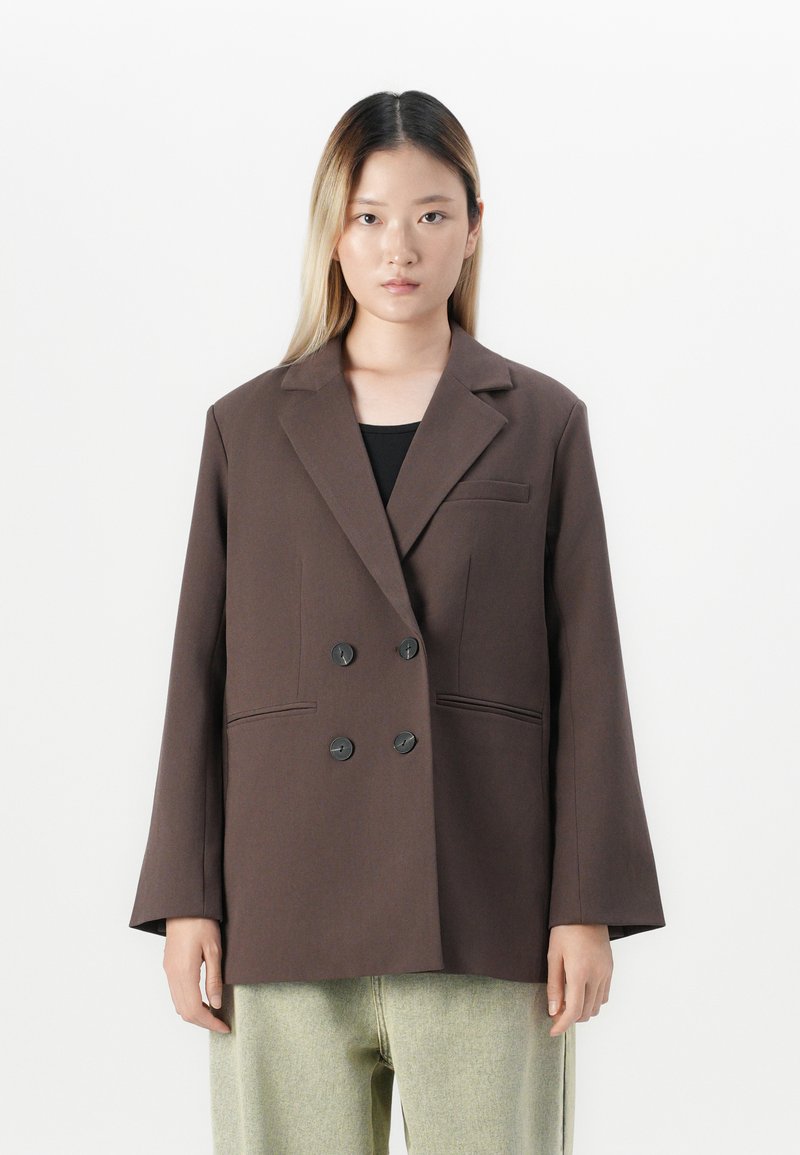 Jeune femme aux cheveux blonds raides portant un blazer oversize marron foncé à double boutonnage et un pantalon léger ample, se tenant devant un fond blanc.