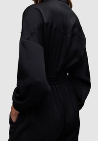 Combinaison noire en tissu lisse, avec une taille ceinturée, des manches larges et des poches. La vue arrière met en évidence des détails de couture minimaux.