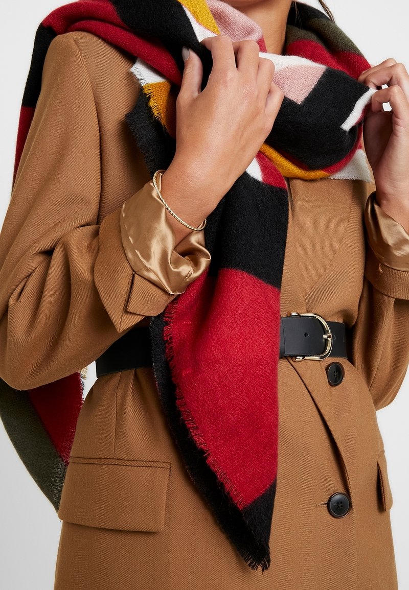 Grande écharpe multicolore en mélange de laine rouge, noir, jaune et rose avec des bords frangés, drapée autour du cou sur une veste marron avec une ceinture.