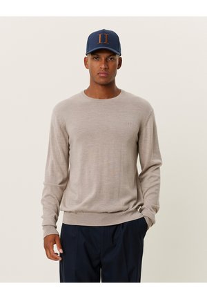 Beige knitted sweater with long sleeves and a round neckline, paired with dark trousers and a navy cap featuring orange accents.