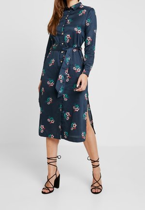 Vrouw die een navyblauwe midi-jurk met lange mouwen en bloemenprint draagt, voorzien van knopen, een tailleceintuur, zijopeningen en zwarte hoge hakken met veter.