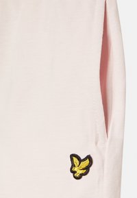 T-shirt rosa claro com mangas curtas, apresentando um pequeno logótipo bordado amarelo e preto de um pássaro no lado inferior esquerdo.