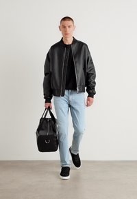 Schwarze Lederbomberjacke, schwarzes T-Shirt, hellblaue Jeans, schwarze Turnschuhe mit weißen Sohlen, trägt eine schwarze Sporttasche, neutraler Hintergrund.