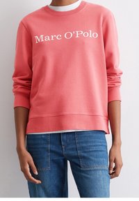 Korale sweter z okrągłym dekoltem i białym napisem "Marc O'Polo". Długie rękawy z ściągaczami, w połączeniu z niebieskimi dżinsami. Gładka faktura.