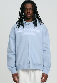 Felpa con zip di colore azzurro chiaro, realizzata in tessuto morbido, caratterizzata da una vestibilità ampia, polsini a coste e il testo ricamato "Low Lights Studios" sul petto.