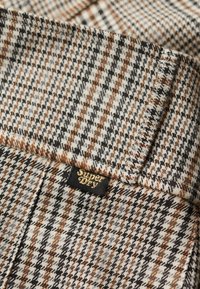 Bruin, grijs en zwart geruit stof met een gestructureerde afwerking. Inclusief een klein zwart label met "Superdry" in gouden letters.