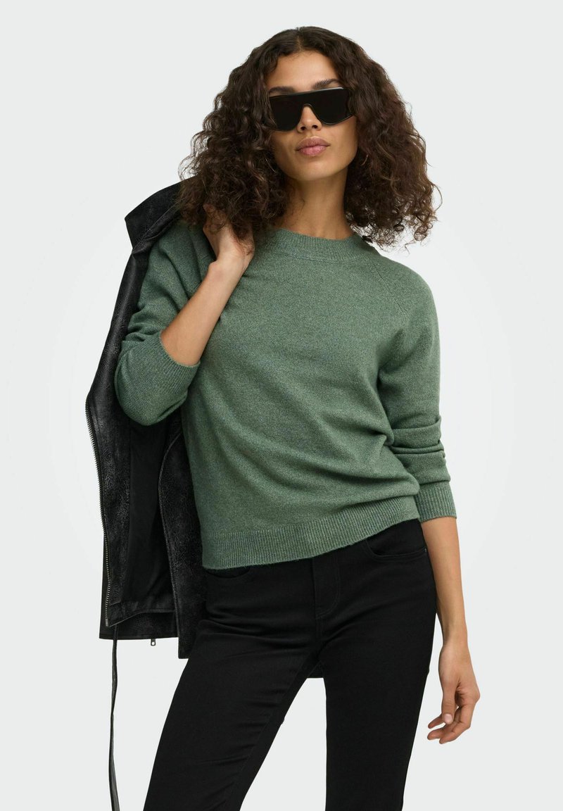 Femme aux cheveux bouclés portant des lunettes de soleil noires, un pull vert, un pantalon noir et tenant une veste noire sur son épaule.