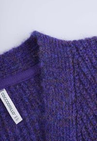 Fioletowy dzianinowy sweter o teksturowanym wykończeniu, z dekoltem w serek i metką oznaczoną "COCCODRILLO". Tkanina zawiera niebieskie i brązowe włókna.
