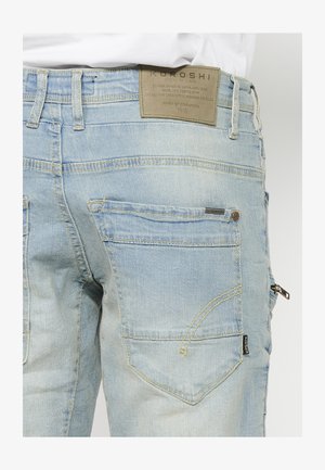 Vaaleansiniset farkut, joissa on vetoketjullinen tasku, kaksi takataskua, keltainen ommel ja nahkalla varustettu etiketti, jossa on bränditietoa vyötäröllä.