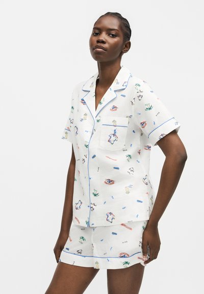 Νεαρή γυναίκα που φοράει λευκή πυτζάμα με κοντά μανίκια, πολύχρωμα σχέδια και μπλε λεπτομέρειες, στέκεται με τα χέρια χαλαρά σε απλό φόντο.