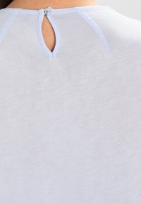 Haut gris clair avec une texture lisse, présentant un détail de dos en trou de serrure et un petit bouton. Couture contrastante le long de l'encolure.