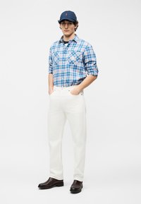 Άνδρας με ναυτικό σκουφί, μπλε καρό πουκάμισο με τυλιγμένα μανίκια, λευκό παντελόνι και σκούρα καφέ μπότες, όρθιος με τα χέρια στις τσέπες.