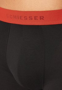 Boxer in jersey nero con elastico rosso in vita, con il nome del marchio "SCHIESSER" in un colore a contrasto, texture liscia.