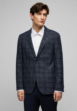 Marineblauer Karo-Blazer aus strukturiertem Material, mit einreihiger Knopfschluss, Reverskragen und zwei vorderen Taschen, kombiniert mit einem weißen Hemd.