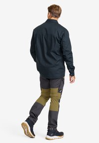 Man som går bort iförd en mörk långärmad skjorta, tvåtonade grå och olivfärgade byxor med knälappar, och mörka vandringsskor med beige sulor.