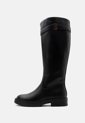 Botte haute en cuir noir avec un talon bas et une sangle décorative avec un emblème en métal près du haut, vue de profil sur fond blanc.