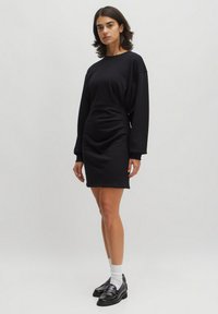 Schwarzes Sweatshirtkleid mit langen Ärmeln, gerafftem Seitendetail und lockerer Passform. Glatte Textur, knielang, kombiniert mit schwarzen Loafers und weißen Socken.