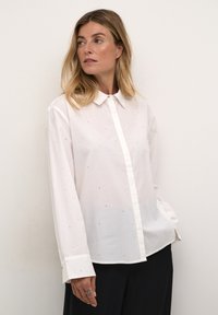 Camisa blanca de botones, hecha de una tela ligera, con sutiles puntos azules, mangas largas y un cuello clásico.