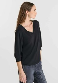 Donna in top nero a V con maniche lunghe fino al gomito e jeans grigi scuri, in piedi di lato contro uno sfondo chiaro e uniforme.