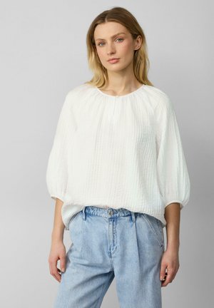 Blouse blanche texturée avec un col rond et un détail de boutons à l'avant, associée à un jean en denim bleu clair taille haute.