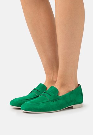 Mocasines de ante verde con una textura suave, que presentan una costura decorativa y un tacón bajo y plano. El diseño es minimalista y elegante.