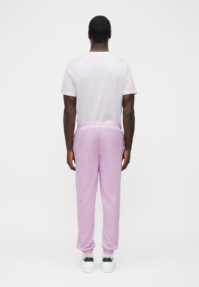 Mężczyzna stojący tyłem, ubrany w białą koszulkę, jasnofioletowe spodnie dresowe, białe skarpetki i białe tenisówki, na jednolitym tle.