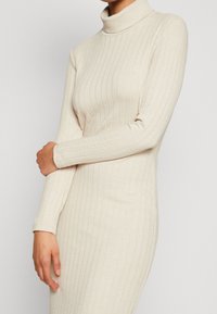 Robe à col roulé à manches longues en tissu côtelé beige clair, présentant une coupe ajustée et des détails texturés le long de la manche et du corps.