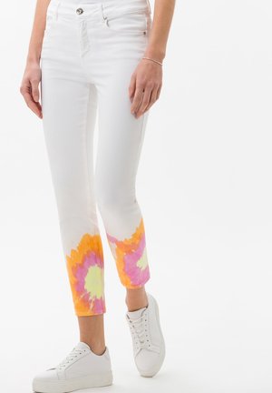 Kobieta w białych skróconych dżinsach z pomarańczowymi, różowymi i żółtymi wzorami tie-dye w okolicach kostek oraz białych tenisówkach.