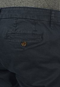 Navyblauwe broek van textuurstof, met een achterzak met een bruine knop en zichtbare stikdetails, gericht op de achteraanzicht.