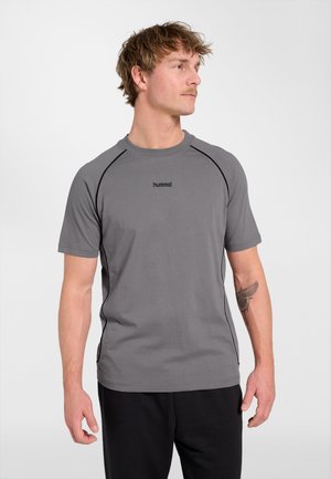 Jonge man met kort haar en snor, draagt een grijs hummel t-shirt en zwarte broek, kijkt naar links, tattoo zichtbaar op linker onderarm.