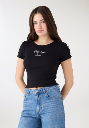 Giovane donna con capelli castani lunghi che indossa una t-shirt nera corta con la scritta "Self Love Club" e jeans azzurri chiari, in piedi davanti a uno sfondo bianco.