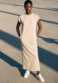 Young person standing on sunlit concrete, wearing a beige sleeveless long dress with pockets and white sneakers, hands in pockets.