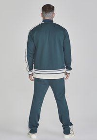 Chándal verde azulado con franjas laterales blancas. Con cuello y puños acanalados, y un ajuste relajado. Fabricado con tejido liso y elástico.
