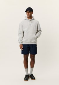 Γκρι φούτερ με κουκούλα και τσέπη καγκουρό, σε συνδυασμό με σκούρο μπλε σορτς. Μαύρο καπέλο και μαύρα παπούτσια, συμπληρώνονται με γκρι κάλτσες. Απλός σχεδιασμός.