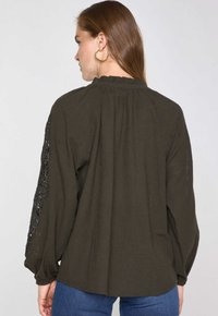 Blusa verde oliva scuro con maniche lunghe a palloncino, con dettagli in pizzo sulle maniche, scollo arruffato e vestibilità ampia.