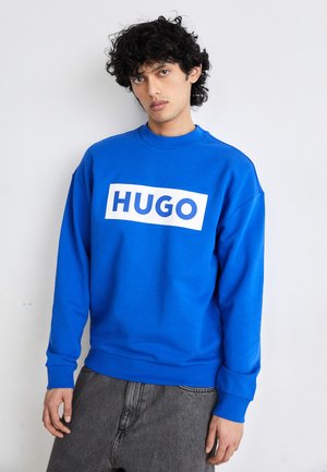 Mėlynas džemperis, pagamintas iš minkštos medžiagos, su dideliu baltu stačiakampiu logotipu, kuriame parašyta "HUGO". Raukšlėtos rankovių ir juosmens dalys.