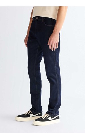 Homme portant un jean slim bleu foncé et des baskets noires et blanches, debout sur un fond clair.