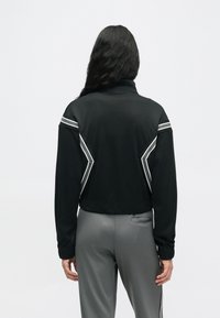 Persona ar gariem melniem matiem, ģērbusies melnā jakā ar baltām svītrām un pelēkās biksēs, stāv uz vienkrāsaina baltā fona, vērsta prom.